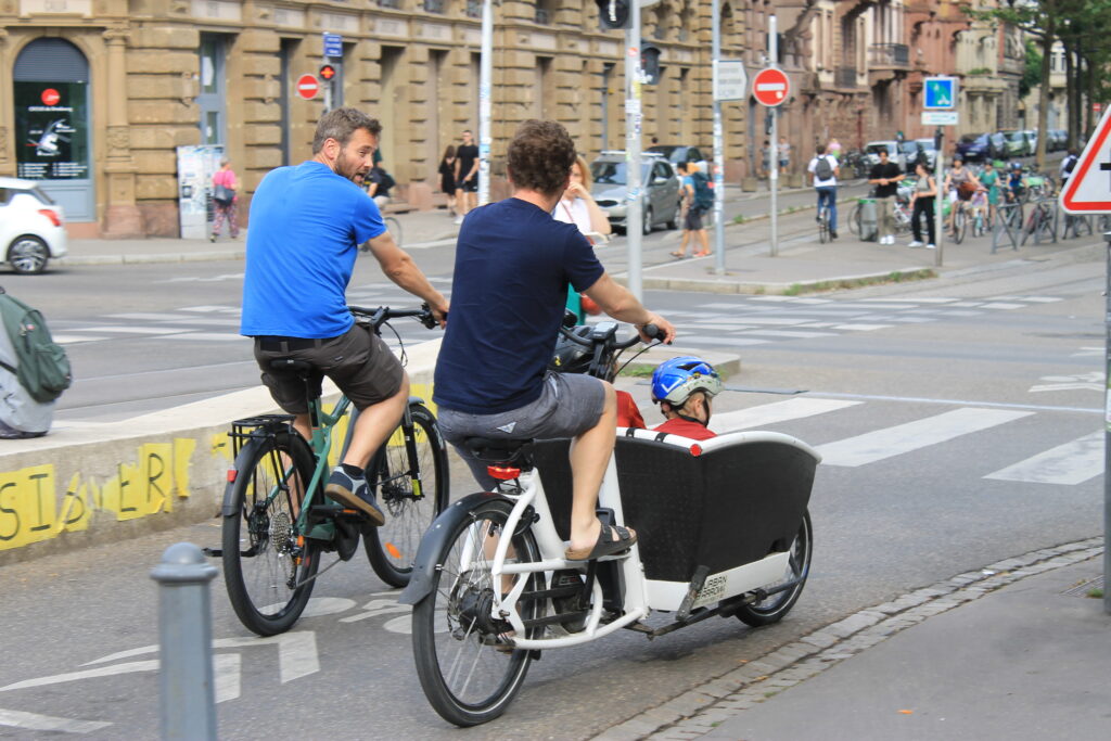 Zwei Radfahrer in Straßburg, davon ein Lastenrad mit einem Kind vorne.