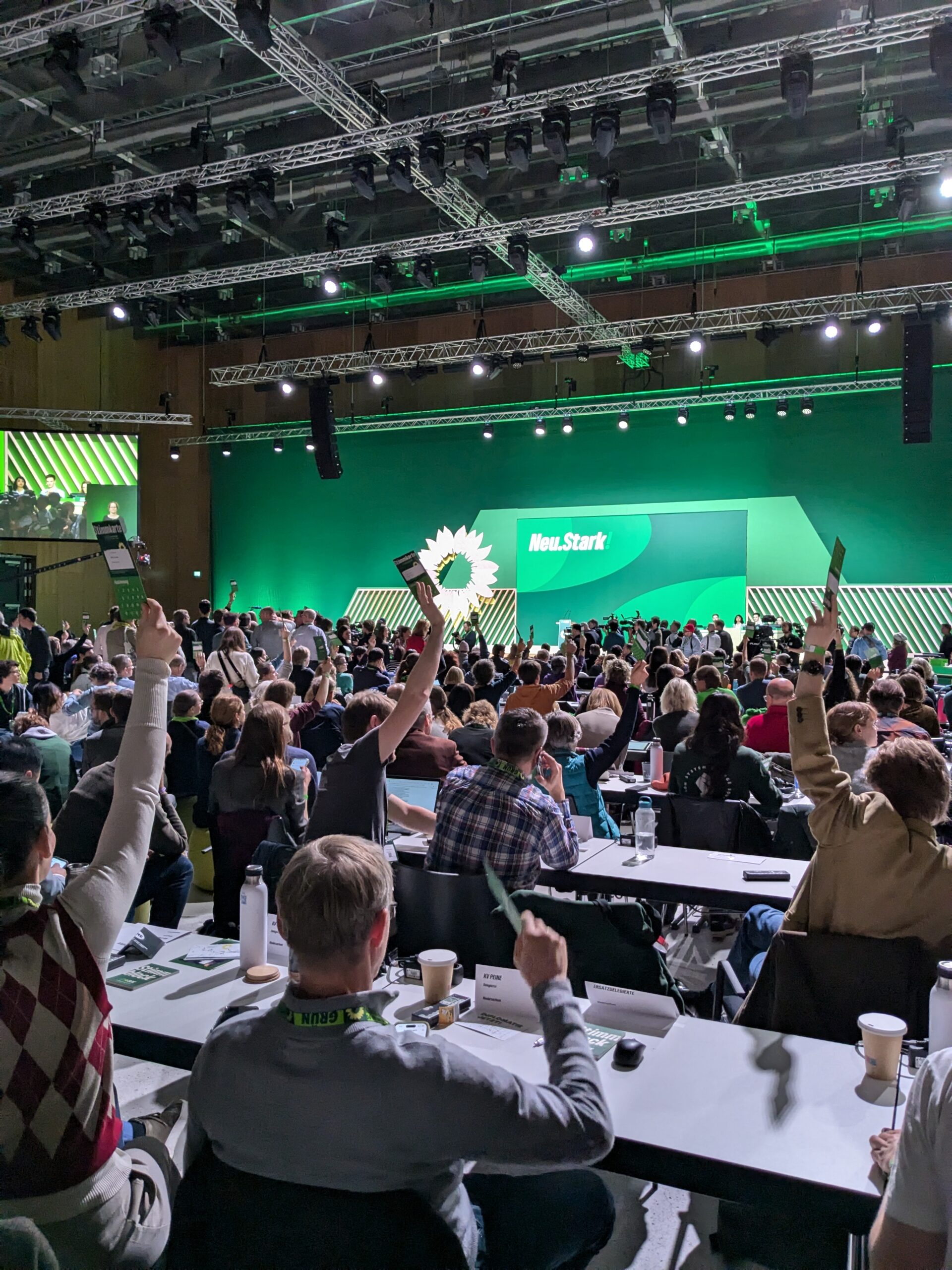 Eine Messehalle mit vielen Menschen an Tischen, im Hintergrund eine grüne Bühne