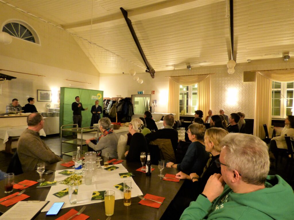 Viele Menschen sitzen an Tischen in einem Saal, im Hintergrund mehrere Personen stehend