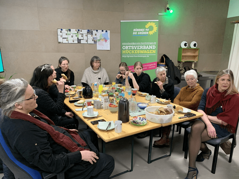Frauen aller Altersstufen sitzen in Hufeisenform um einen Holztisch in der Geschäöftsstelle. Der Tisch ist zum Frühstück üppig gedeckt. An der Wand das ein oder andere Plakat und Infoblatt der Partei.