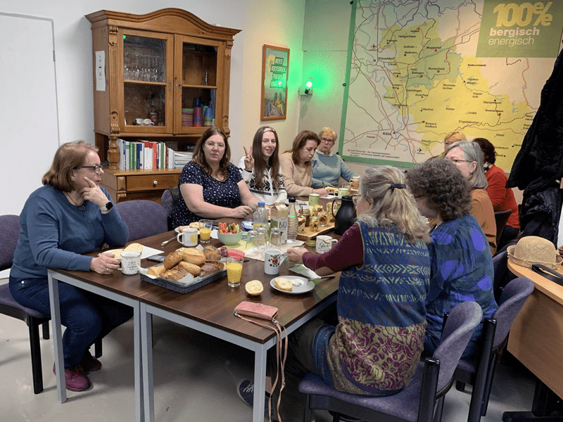 Frauen aller Altersstufen sitzen in Hufeisenform um einen Holztisch in der Geschäöftsstelle. Der Tisch ist zum Frühstück üppig gedeckt. An der Wand stzeht eine Landhausstil-Vitrine, rechts an der hinteren Wand eine Landkarte des Bergischen prominent im Bild, darüber steht '100% Bergisch Energisch'