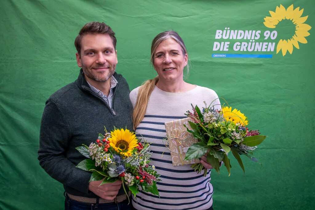 Dr. Julian Münster und Sylvia Berghoff, die einstimmig gewählten Landtagskandidat\*innen der GRÜNEN Oberberg für die Landtagswahl 2027, mit Blumensträußen vor einer grünen Parteiwand mit Sonnenblumenlogo.